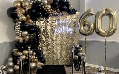 60th birthday setup with the vanilla gold sequin wall, balloons, neon sign, helium numbers, personalised cake stand & black carpet