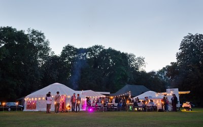  A party with yurts, wood fired pizza vans, decorative outdoor lamps, and groups of people. 