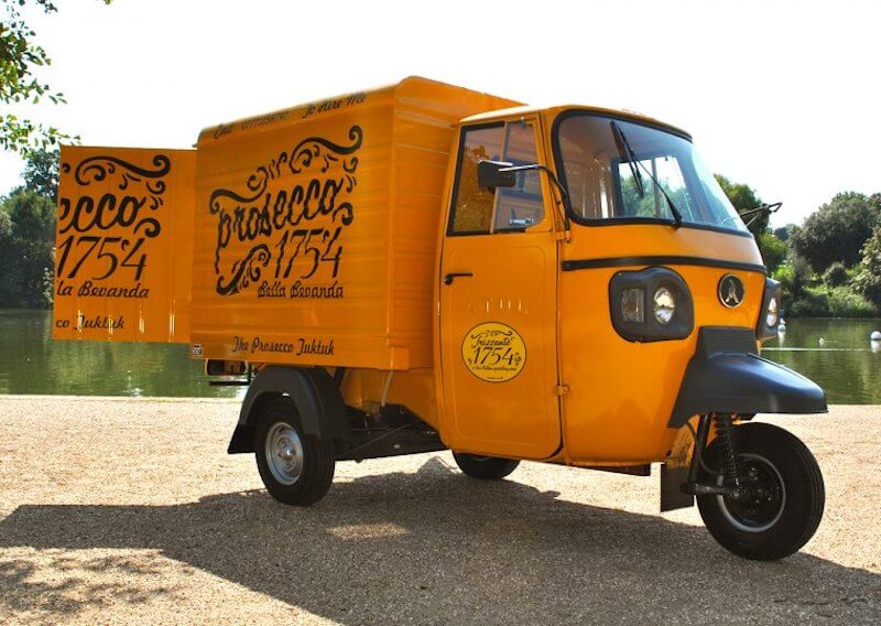 The eye-catching yellow Prosecco Tuk Tuk van in front of a lake