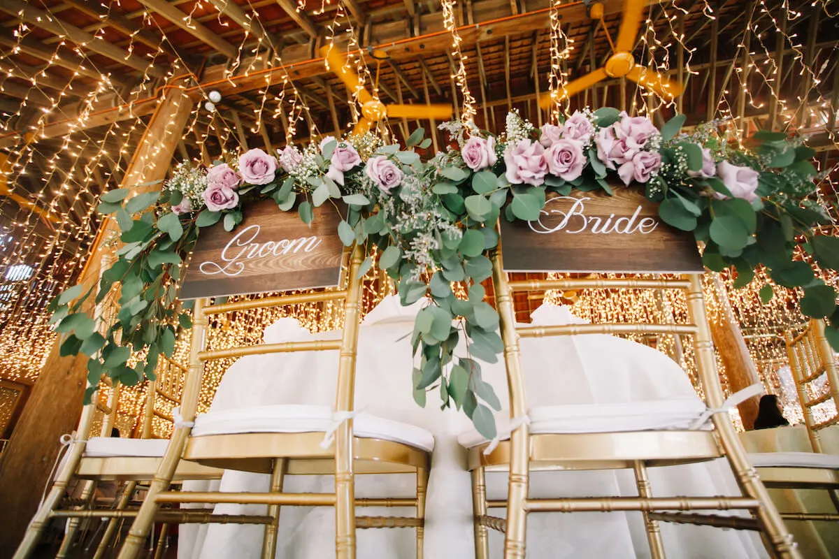 Decorated bride and groom chairs within a barn covered in lights