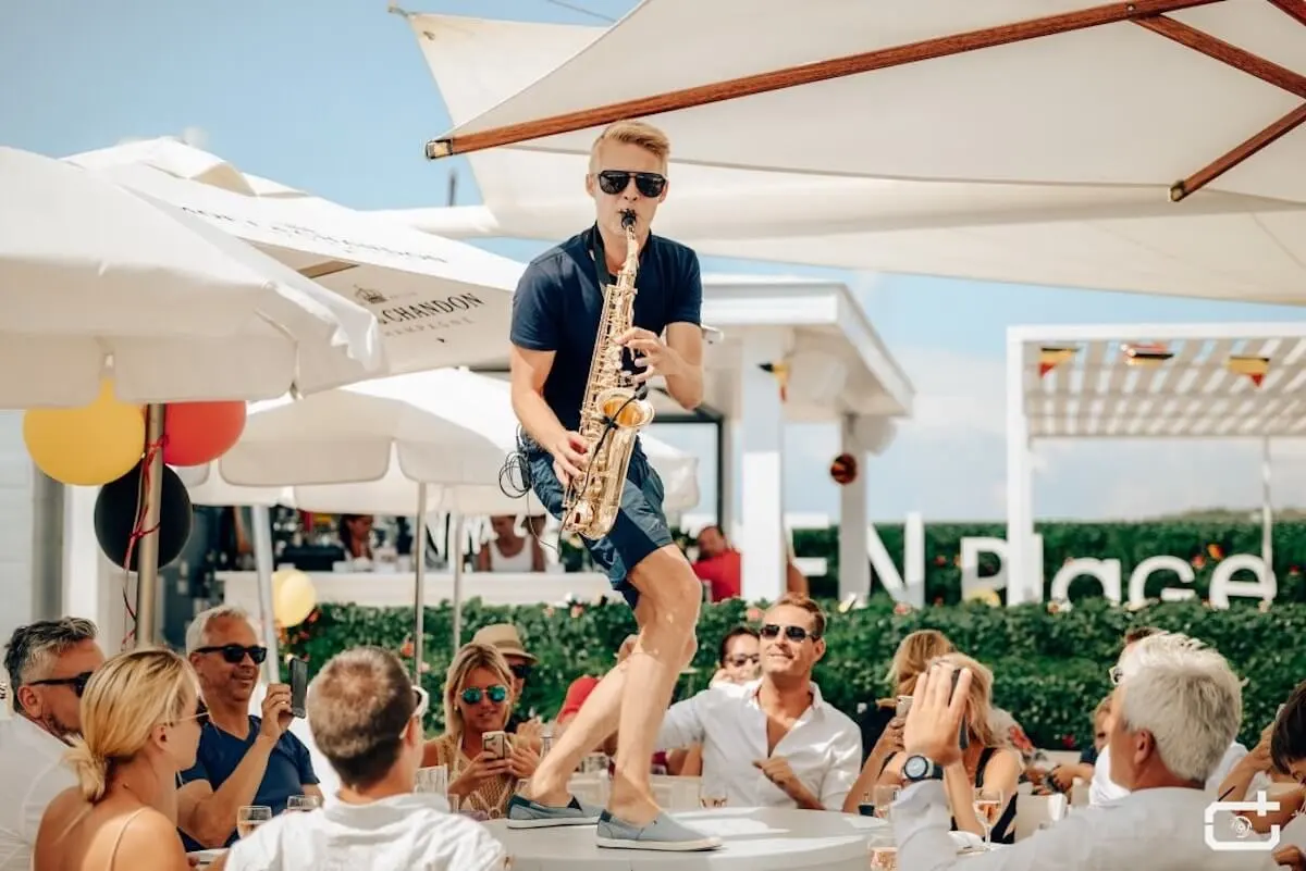 Image of performer playing the saxophone whilst standing on a table surrounded by an audience